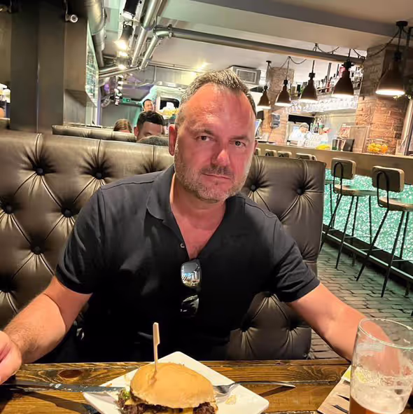 Mężczyzna siedzący przy stoliku w restauracji z burgerem i napojem, ubrany w ciemną koszulkę polo i okulary przeciwsłoneczne zawieszone na kołnierzu.