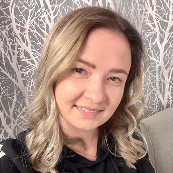 Uśmiechnięta kobieta o blond falowanych włosach, ubrana w czarny top, przed szaro-białą ścianą z wzorem gałęzi.
